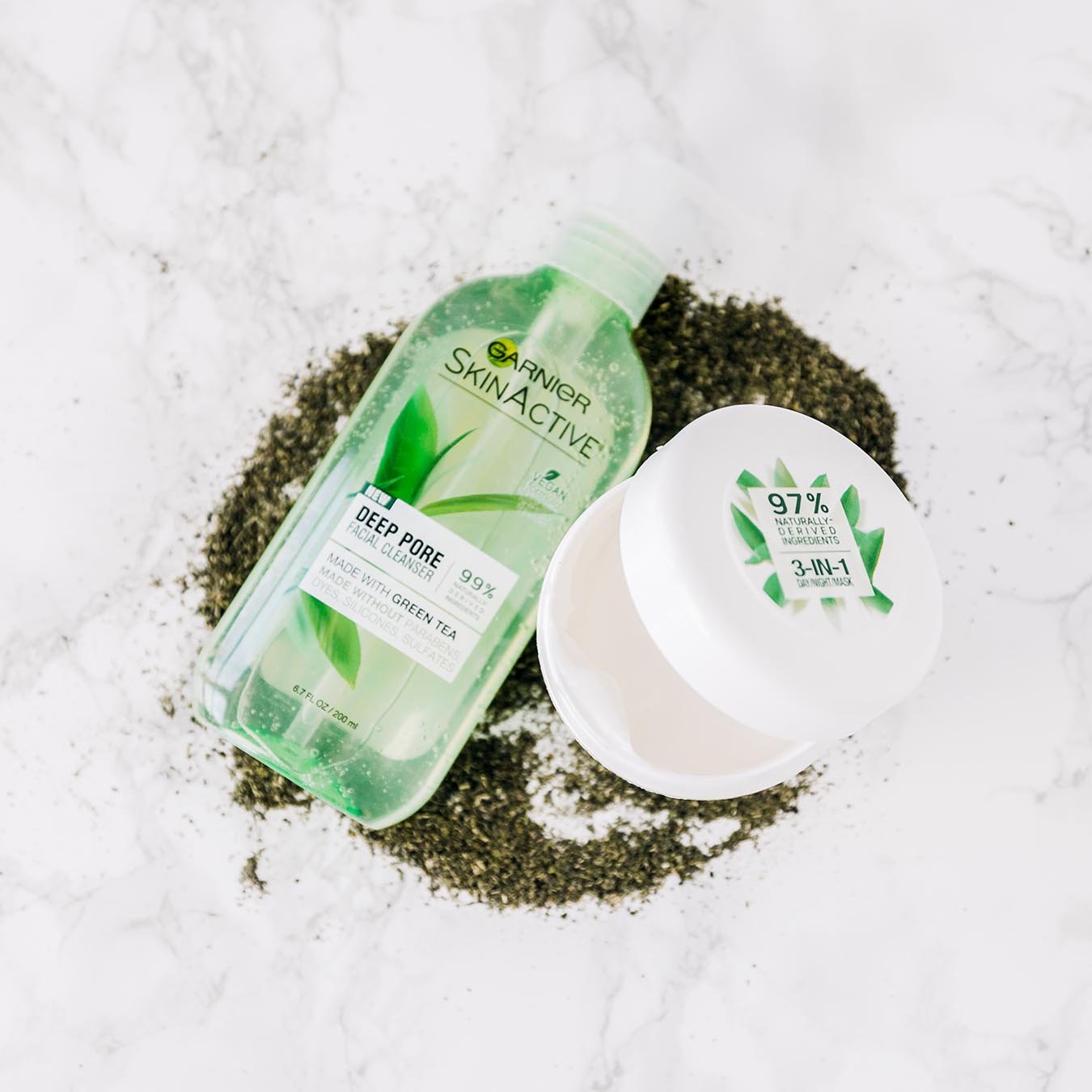Garnier SkinActive 3-in-1 Day/Night Mask ajar with Green Tea and SkinActive Deep Pore Cleanser sitting in a pile of powdered tea leaves on white marble.